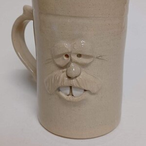 K&ouml;nnte beinhalten: Beigefarbene Keramiktasse mit einem geformten Gesichtsdesign. Die Tasse hat einen Henkel und ein detailliertes Gesicht mit Schnurrbart, Nase, Augen und Z&auml;hnen. Die Tasse hat eine strukturierte Oberfl&auml;che und eine zylindrische Form.
