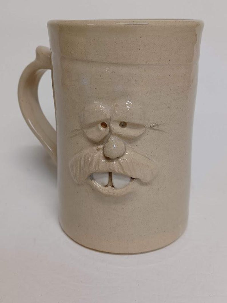 K&ouml;nnte beinhalten: Beigefarbene Keramiktasse mit einem geformten Gesichtsdesign. Die Tasse hat einen Henkel und ein detailliertes Gesicht mit Schnurrbart, Nase, Augen und Z&auml;hnen. Die Tasse hat eine strukturierte Oberfl&auml;che und eine zylindrische Form.