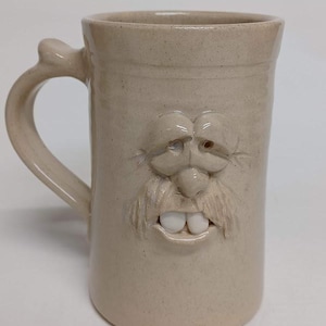K&ouml;nnte beinhalten: Beigefarbene Keramiktasse mit einem geformten Gesichtsdesign. Die Tasse hat einen Henkel und ein detailliertes Gesicht mit Schnurrbart, Nase, Augen und Z&auml;hnen. Die Tasse ist etwa 15 cm hoch.
