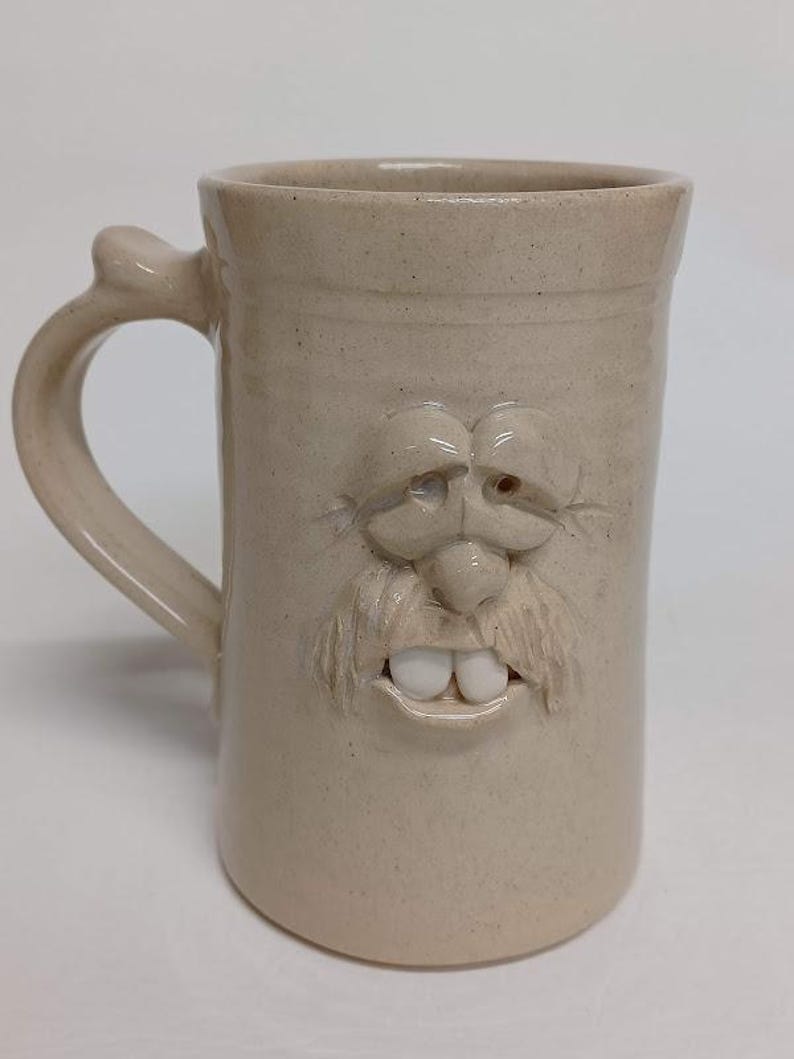 K&ouml;nnte beinhalten: Beigefarbene Keramiktasse mit einem geformten Gesichtsdesign. Die Tasse hat einen Henkel und ein detailliertes Gesicht mit Schnurrbart, Nase, Augen und Z&auml;hnen. Die Tasse ist etwa 15 cm hoch.