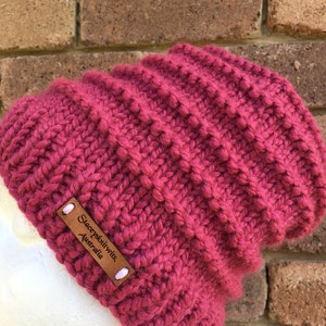 Pode incluir: Um gorro de malha rosa escuro com uma etiqueta de couro marrom que diz "Stacey's Knitwits, Australia".