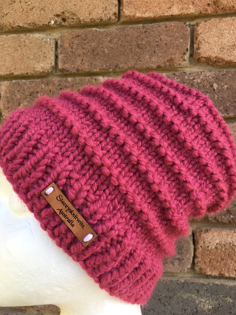 Pode incluir: Um gorro de malha rosa escuro com uma etiqueta de couro marrom que diz "Stacey's Knitwits, Australia".