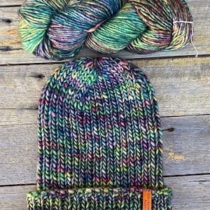 May include: A hand-knitted beanie and a skein of yarn in vibrant colors, including green, purple, and yellow. The beanie has a folded brim and a small tag. The yarn is a thick, textured wool, perfect for knitting or crocheting projects.