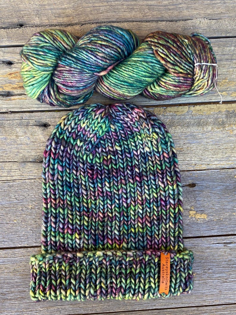 May include: A hand-knitted beanie and a skein of yarn in vibrant colors, including green, purple, and yellow. The beanie has a folded brim and a small tag. The yarn is a thick, textured wool, perfect for knitting or crocheting projects.