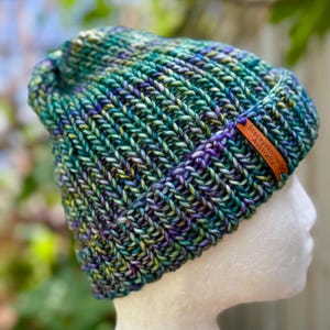 May include: A hand-knitted beanie in shades of teal, green, purple, and yellow. The hat features a ribbed brim and a tag that reads "Staceysknitwitz Australia."