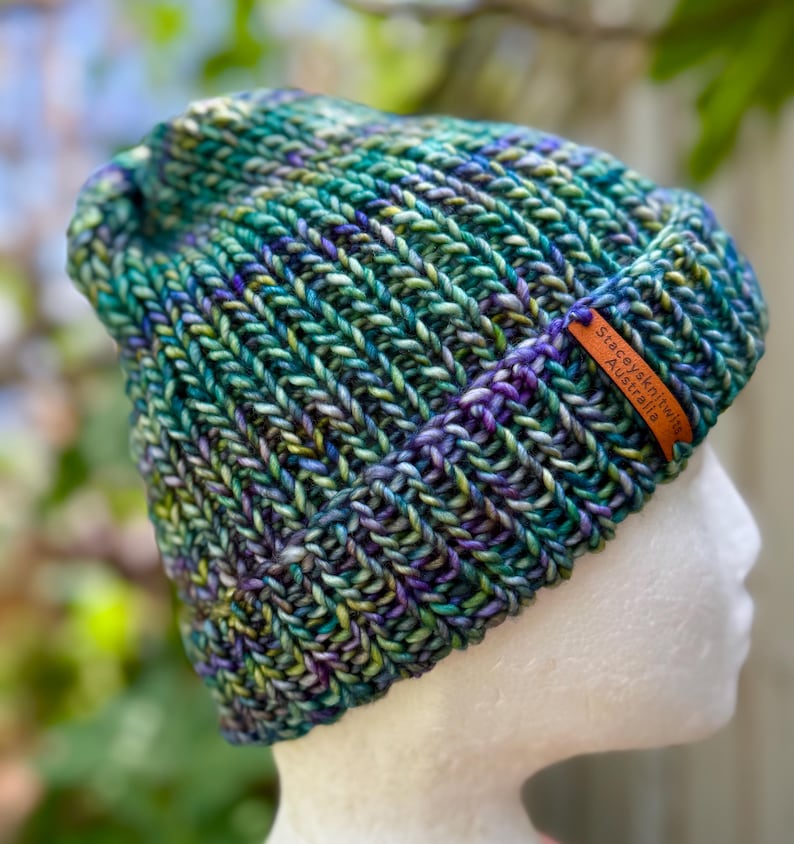 May include: A hand-knitted beanie in shades of teal, green, purple, and yellow. The hat features a ribbed brim and a tag that reads "Staceysknitwitz Australia."