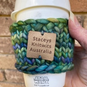 Op de afbeelding: Een groene, blauwe en gele gebreide koffiebekerwarmer met een bruin leren label waarop "Stacey's Knitwits Australia" staat. De warmer zit op een witte koffiebeker met het Star Wars-logo.