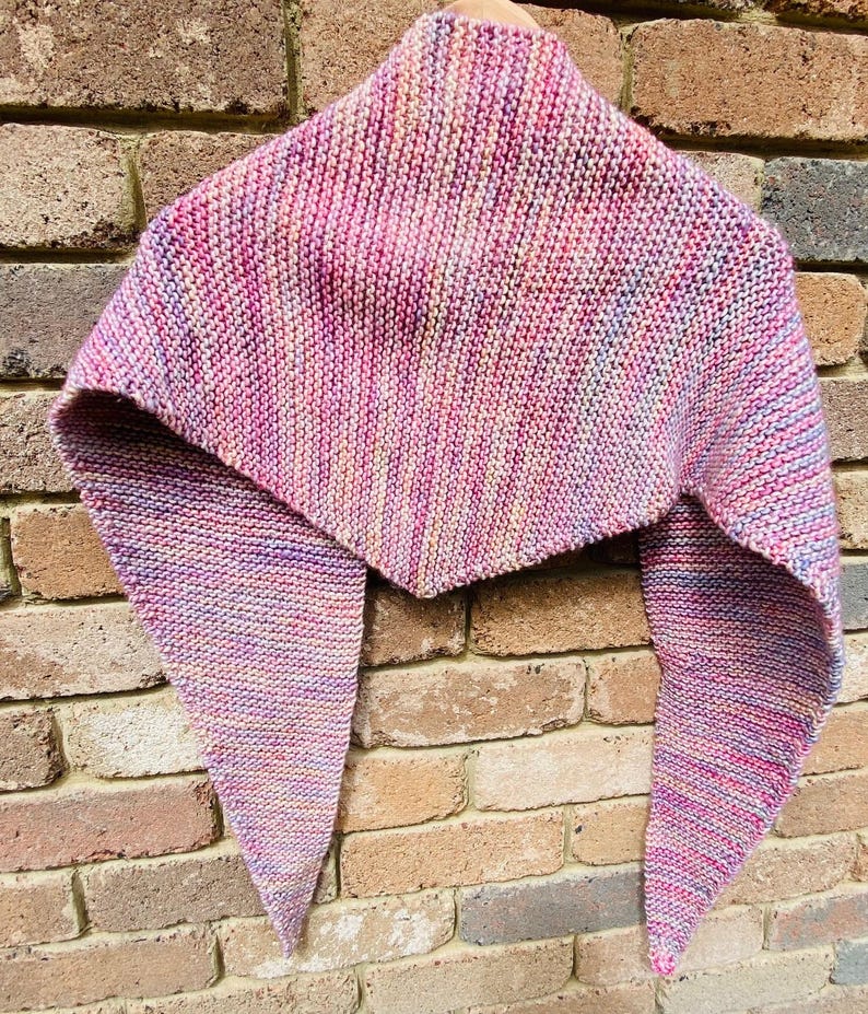 May include: A triangular knit shawl featuring a variegated color palette of pink, purple, orange, and blue tones. The garment is crafted with a consistent garter stitch texture, creating a soft, textured appearance with pointed ends and a wide center.