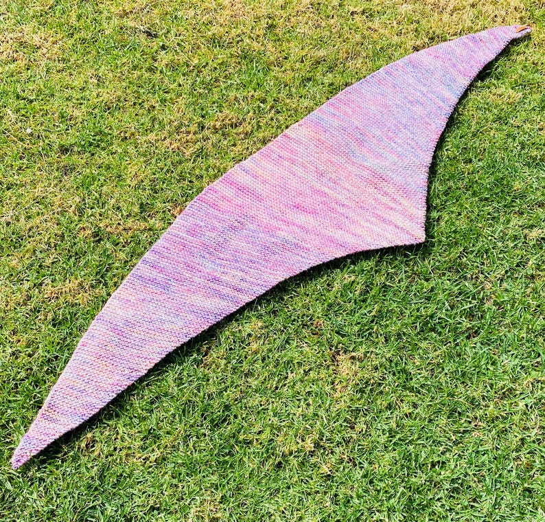 May include: A triangular knitted shawl featuring a variegated color palette of soft pink, lavender, and subtle orange hues. The textile displays a consistent garter stitch pattern throughout, with a tapered shape that extends to pointed ends.