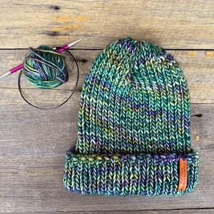 Könnte beinhalten: Eine handgestrickte Mütze in Grün-, Blau- und Gelbtönen mit umgeschlagenem Saum. Ein Knäuel Wolle und Stricknadeln liegen neben der Mütze. Ein kleines orangefarbenes Etikett am Saum trägt die Aufschrift "Staceykatwits Australia."