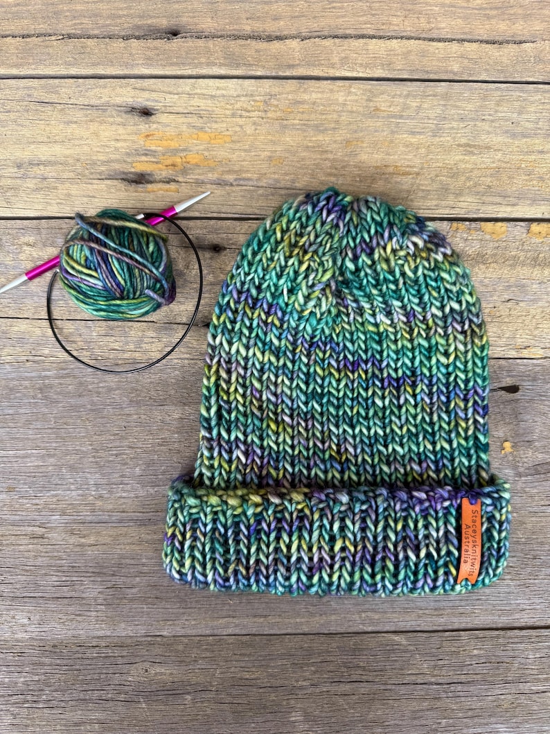May include: A hand-knitted beanie in shades of green, blue, and yellow, with a folded brim. A ball of yarn and knitting needles are next to the hat. A small orange tag on the brim reads "Staceykatwits Australia."