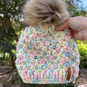 May include: A colorful knitted beanie with a fluffy brown pom pom. The beanie is made with a chunky yarn in shades of pink, blue, yellow, and white.
