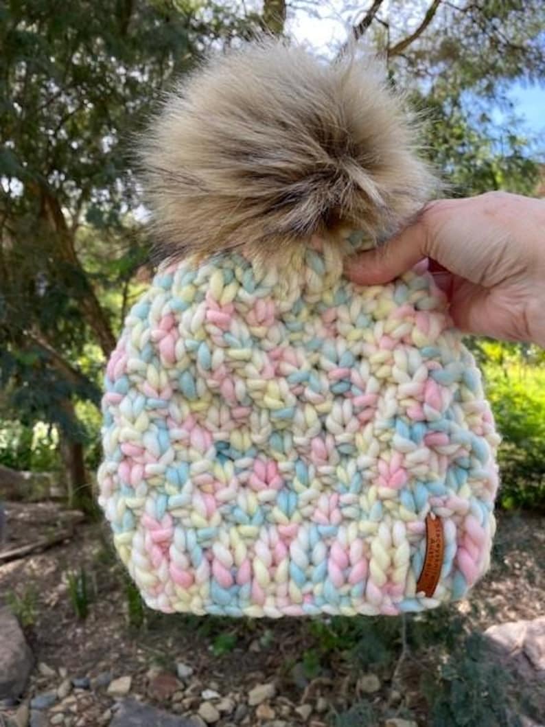 May include: A colorful knitted beanie with a fluffy brown pom pom. The beanie is made with a chunky yarn in shades of pink, blue, yellow, and white.