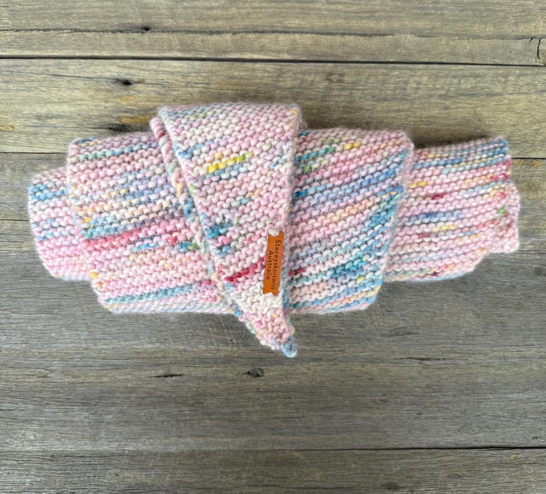 May include: A hand-knitted scarf featuring a variegated yarn pattern with soft pastel pink, blue, yellow, and white flecks. The scarf is folded in a layered arrangement and includes a small, rectangular brown leather tag labeled StaceyKnits Australia.