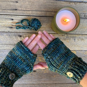 Könnte beinhalten: Ein Paar tealgrüne Fingerlose Handschuhe mit einem Knopf an jeder Manschette. Die Handschuhe sind mit einem strukturierten Stich gestrickt und aus einem Garn hergestellt, das eine subtile Farbvariation aufweist.