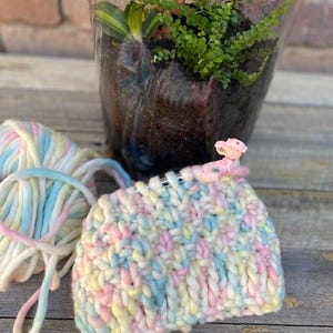 May include: A close-up of a colorful yarn project in progress. The yarn is a mix of pink, blue, and yellow. The project is being worked on with knitting needles and a stitch marker.