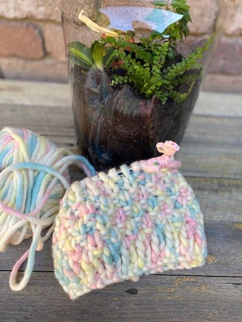 May include: A close-up of a colorful yarn project in progress. The yarn is a mix of pink, blue, and yellow. The project is being worked on with knitting needles and a stitch marker.