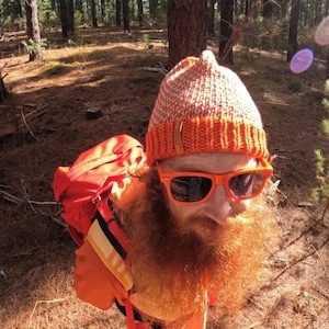 May include: A person wearing a bright orange backpack, a knitted orange and white beanie, and orange sunglasses. The person has a long, red beard and is standing in a forest.