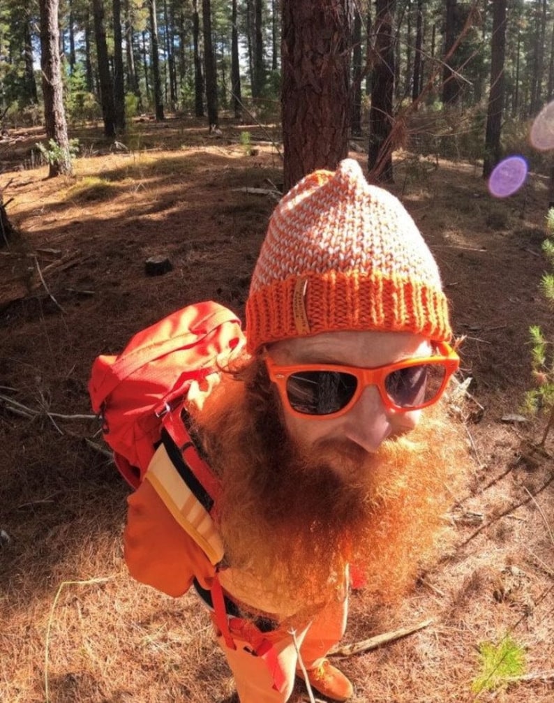 May include: A person wearing a bright orange backpack, a knitted orange and white beanie, and orange sunglasses. The person has a long, red beard and is standing in a forest.