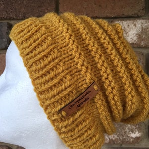 Pode incluir: Um gorro de malha amarelo mostarda com uma etiqueta de couro marrom que diz "Stacey's Knitwits, Australia".