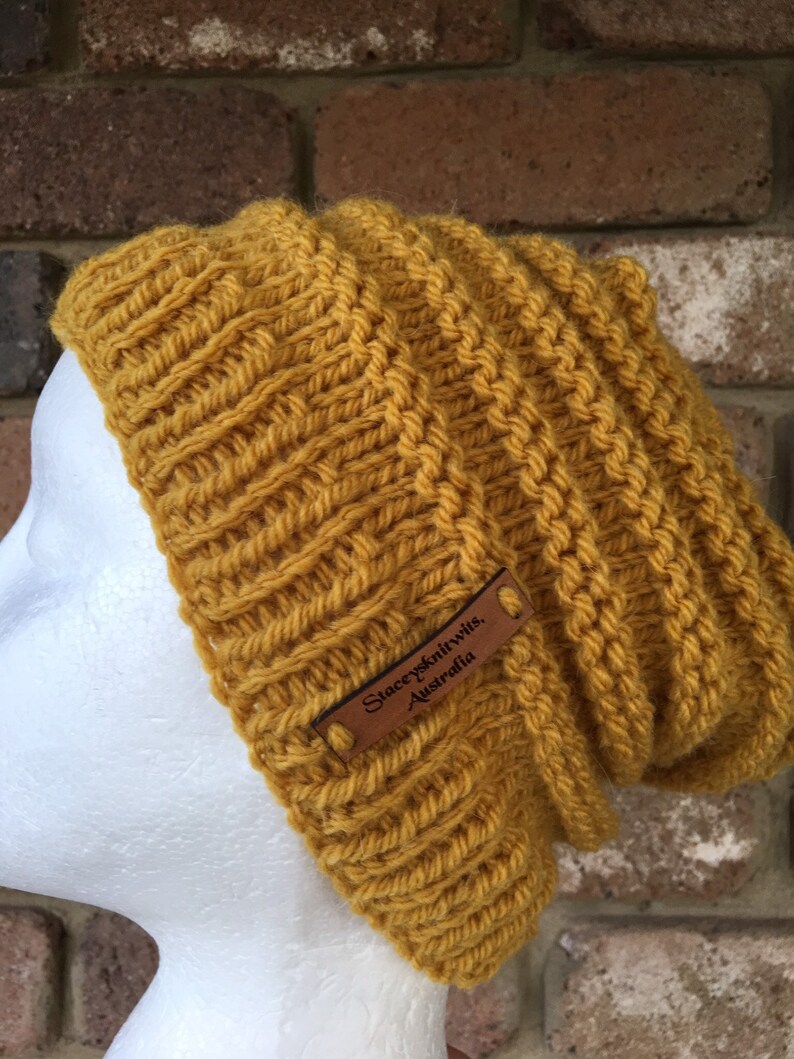 Pode incluir: Um gorro de malha amarelo mostarda com uma etiqueta de couro marrom que diz "Stacey's Knitwits, Australia".