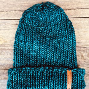 May include: A teal knit beanie with a folded brim. The hat is made of thick yarn and has a textured appearance. A small brown tag with the text "Staceysknits Australia" is attached to the brim.