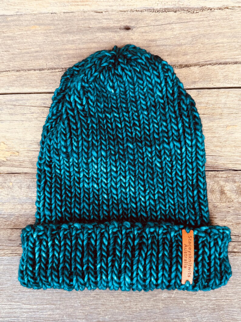 May include: A teal knit beanie with a folded brim. The hat is made of thick yarn and has a textured appearance. A small brown tag with the text "Staceysknits Australia" is attached to the brim.