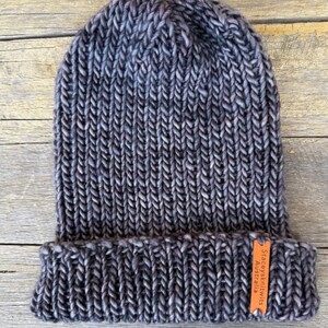 May include: A hand-knitted beanie in shades of gray and blue, with a folded brim. The hat has a ribbed texture and a small orange tag that reads "Staceysknits Australia". The beanie is made of thick yarn.