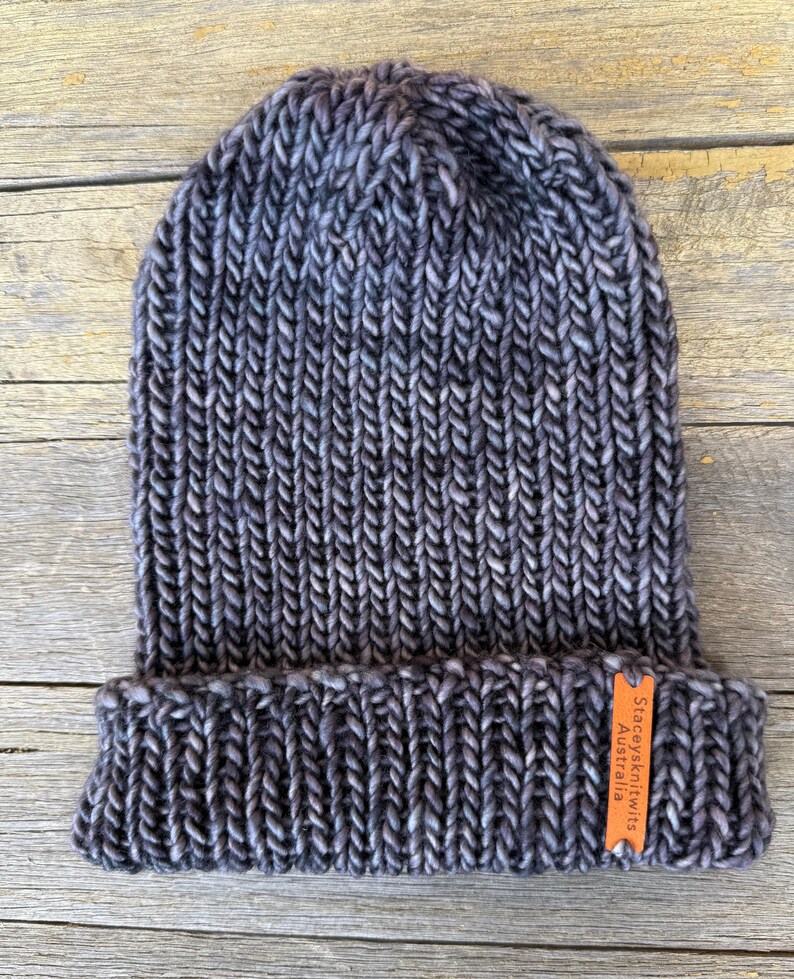 May include: A hand-knitted beanie in shades of gray and blue, with a folded brim. The hat has a ribbed texture and a small orange tag that reads "Staceysknits Australia". The beanie is made of thick yarn.