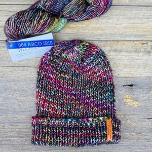 May include: A hand-knitted beanie hat and a skein of yarn in a vibrant color palette of purple, pink, green, and yellow. The hat has a folded brim and a small tag. The yarn is labeled "866 Arco Iris".