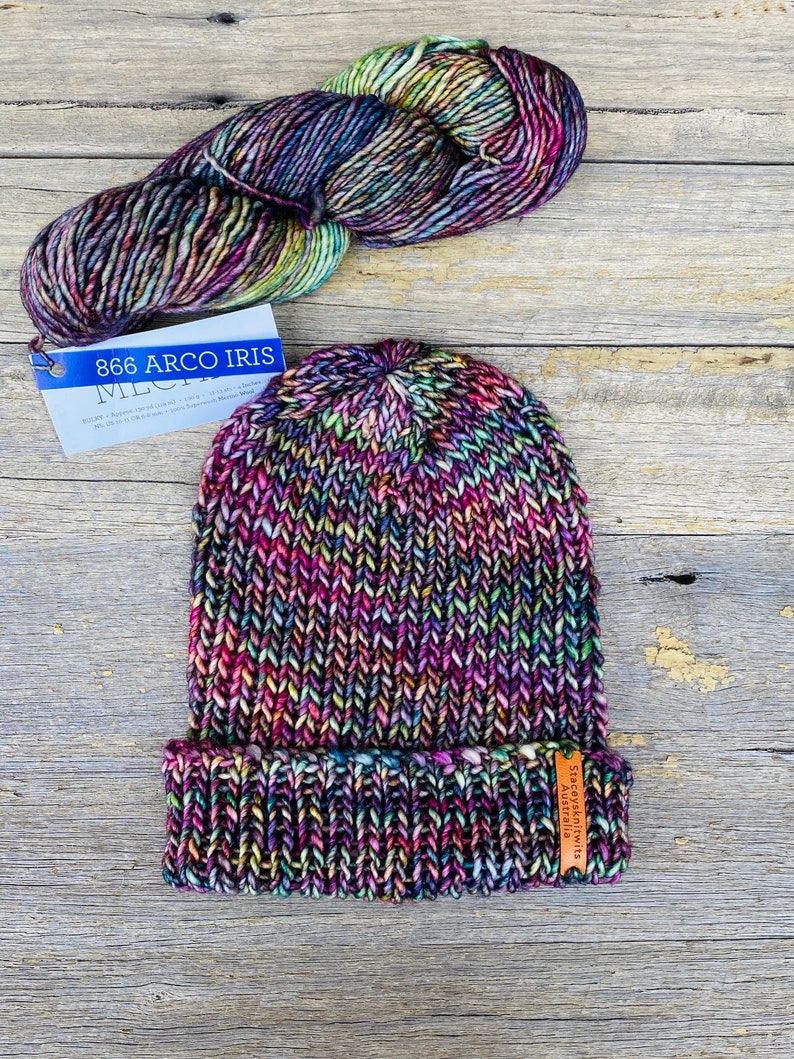May include: A hand-knitted beanie hat and a skein of yarn in a vibrant color palette of purple, pink, green, and yellow. The hat has a folded brim and a small tag. The yarn is labeled "866 Arco Iris".