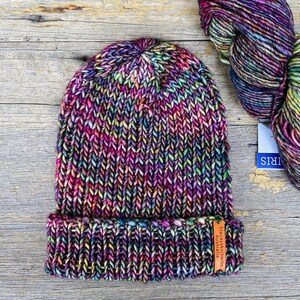 May include: A hand-knitted beanie in a vibrant mix of colors, including purple, pink, green, and yellow. The hat has a folded brim and a tag that reads "Staceysknits Australia". Beside the hat is a skein of matching yarn.