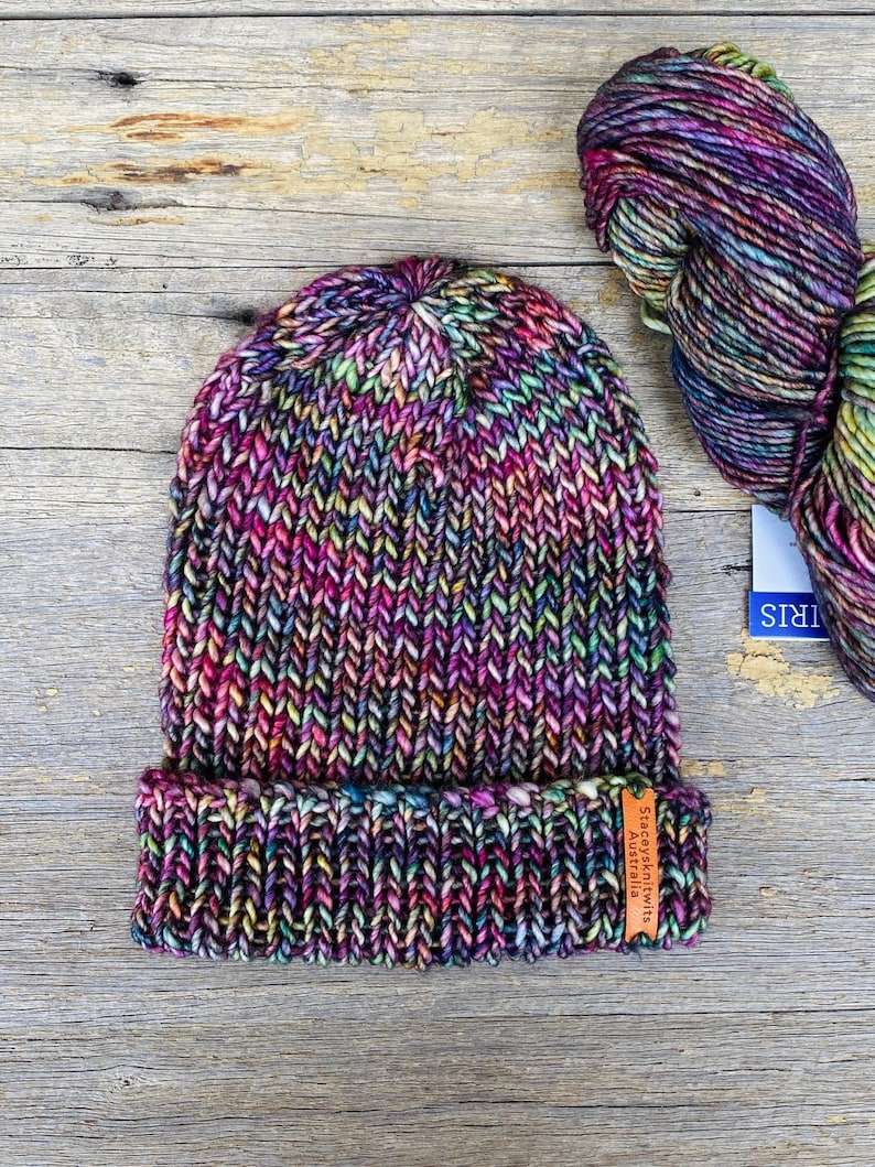 May include: A hand-knitted beanie in a vibrant mix of colors, including purple, pink, green, and yellow. The hat has a folded brim and a tag that reads "Staceysknits Australia". Beside the hat is a skein of matching yarn.