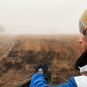May include: A person wearing a yellow and gray knit beanie, a black scarf, and a white jacket with a blue sleeve, looks out at a foggy landscape. The person is sitting on a wooden bench with their hand resting on the bench. The landscape is covered in tall grass and a dirt path leads into the distance.