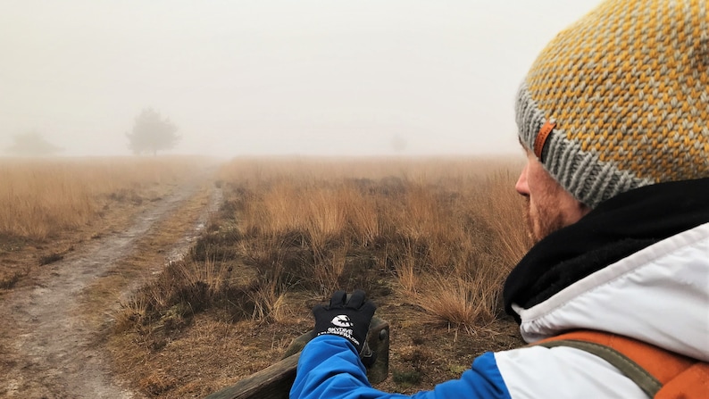 May include: A person wearing a yellow and gray knit beanie, a black scarf, and a white jacket with a blue sleeve, looks out at a foggy landscape. The person is sitting on a wooden bench with their hand resting on the bench. The landscape is covered in tall grass and a dirt path leads into the distance.