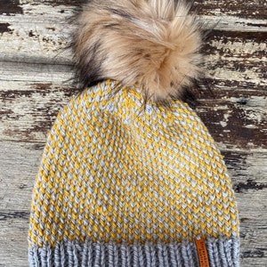 May include: A yellow and gray knitted beanie with a brown faux fur pom pom. The beanie has a gray ribbed cuff and a brown leather tag that says "Seafolly Knits Australia".