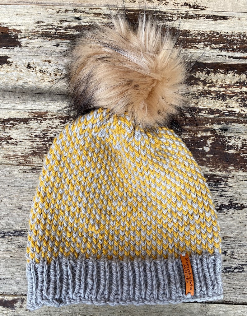 May include: A yellow and gray knitted beanie with a brown faux fur pom pom. The beanie has a gray ribbed cuff and a brown leather tag that says "Seafolly Knits Australia".