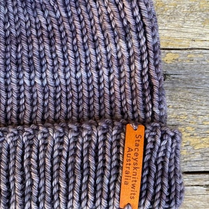 May include: A hand-knitted beanie in shades of gray and purple, with a ribbed texture. A small, brown leather tag is attached, bearing the text "Staceysknitwits Australia". The hat is made of thick yarn.