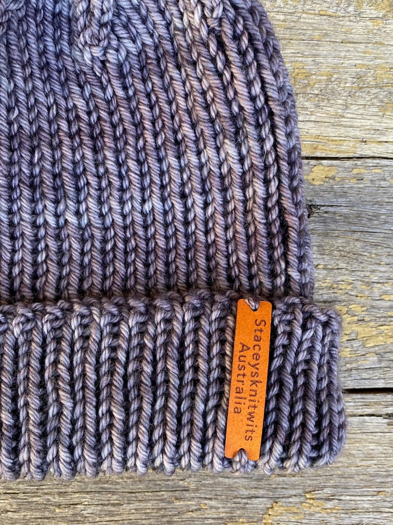 May include: A hand-knitted beanie in shades of gray and purple, with a ribbed texture. A small, brown leather tag is attached, bearing the text "Staceysknitwits Australia". The hat is made of thick yarn.