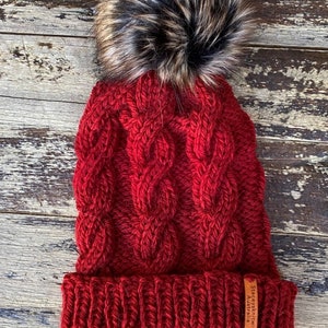 May include: Red knitted beanie with a large brown and black faux fur pom pom. The beanie has a cable knit pattern and a brown leather label that reads "Stacey's Knits Australia".