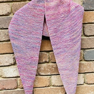 May include: A triangular knit shawl featuring a variegated color palette of pink, purple, blue, and soft orange tones. The garment is crafted with a consistent garter stitch texture and includes a small brown leather tag attached to one corner.