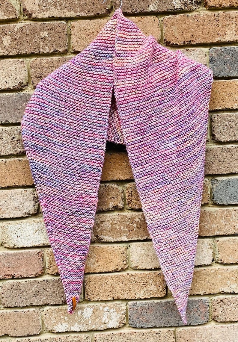 May include: A triangular knit shawl featuring a variegated color palette of pink, purple, blue, and soft orange tones. The garment is crafted with a consistent garter stitch texture and includes a small brown leather tag attached to one corner.