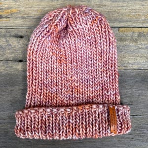 May include: A hand-knitted beanie in shades of pink, peach, and white. The hat features a folded brim and a chunky knit texture. A small leather tag with text is attached to the brim. The hat is displayed on a wooden surface.