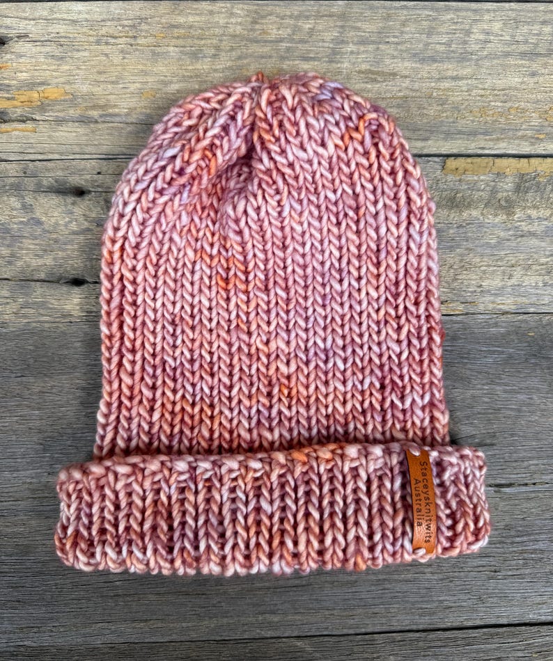 May include: A hand-knitted beanie in shades of pink, peach, and white. The hat features a folded brim and a chunky knit texture. A small leather tag with text is attached to the brim. The hat is displayed on a wooden surface.
