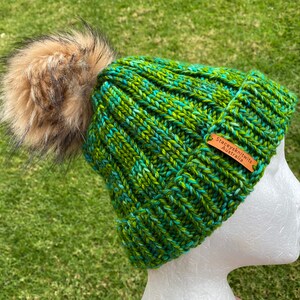 May include: Green and blue knitted beanie with a brown faux fur pom pom. The beanie has a brown leather tag that says "Stacey's Knits Australia".