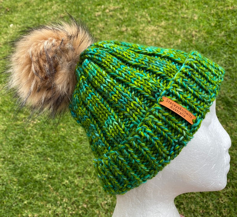 May include: Green and blue knitted beanie with a brown faux fur pom pom. The beanie has a brown leather tag that says "Stacey's Knits Australia".