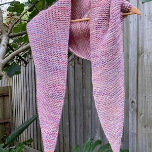 May include: A triangular knit scarf featuring a variegated color palette of pink, purple, and orange yarn. The scarf is constructed with a garter stitch pattern and includes a small, rectangular orange leather-like tag attached to one corner.