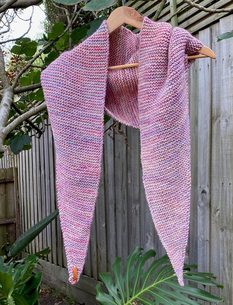 May include: A triangular knit scarf featuring a variegated color palette of pink, purple, and orange yarn. The scarf is constructed with a garter stitch pattern and includes a small, rectangular orange leather-like tag attached to one corner.
