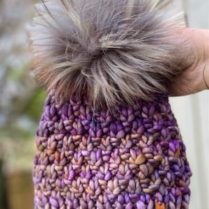 May include: Purple and brown knitted beanie with a fluffy grey pom pom. The beanie has a woven label that reads "Stacey Knitwear Australia".