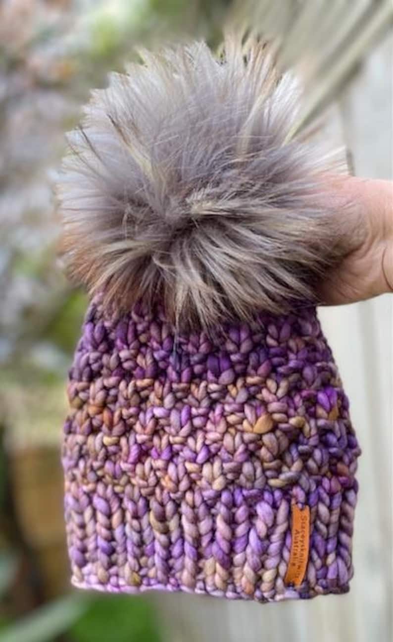 May include: Purple and brown knitted beanie with a fluffy grey pom pom. The beanie has a woven label that reads "Stacey Knitwear Australia".